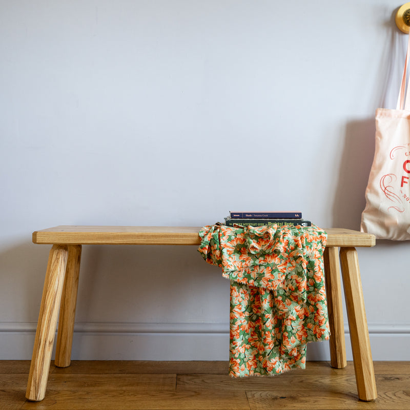 Bench against a wall between fabric and two books placed on top. A tote bag is hanging up to the right of the bench