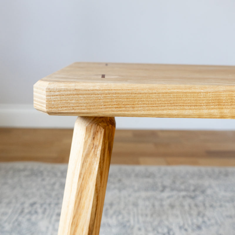 Close up of an ash bench with octagonal legs. An off-white wall is in the background. The bench is on a patterned rug.