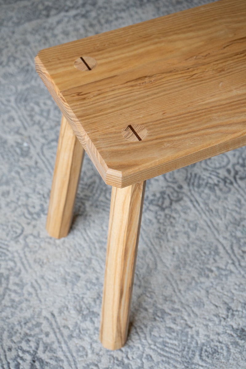 Top, angled view of an ash bench with octagonal legs on a patterned rug. The focus is on the wood grain and joinery detail on the top surface.