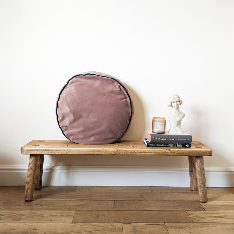 Pine bench finished in Medium Brown against an off white wall. There is a round, pink cushion on the bench along with two books, a candle and a ornament.