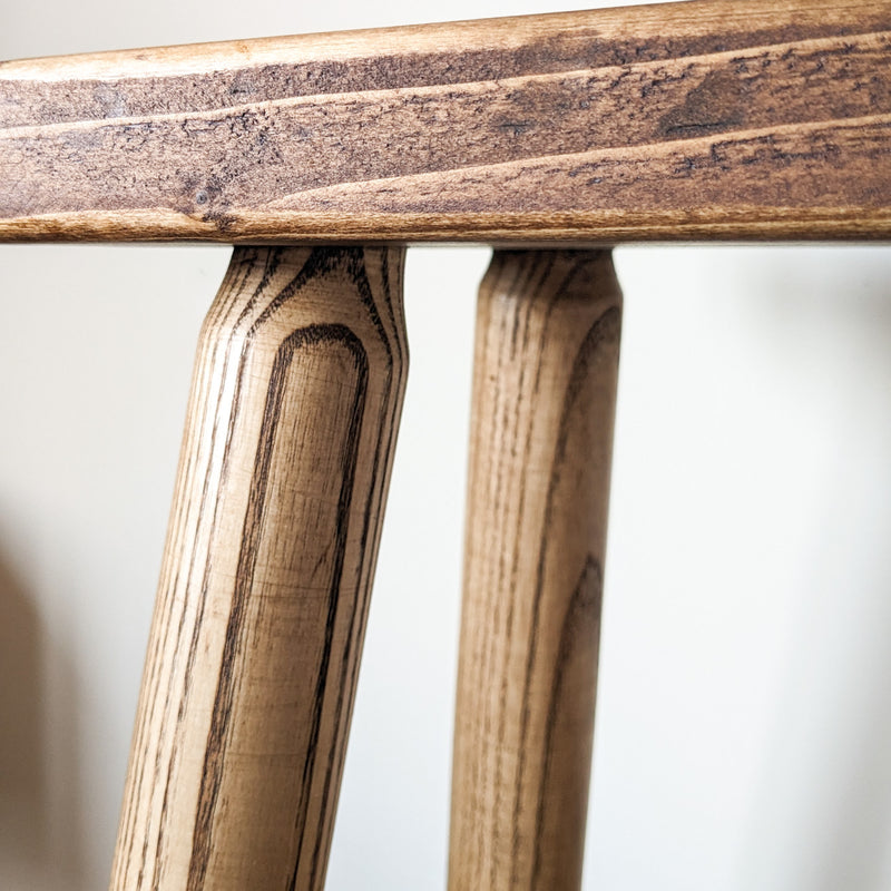Close up of a pine bench finished a Warm, Dark Brown. The focus is on the chunky legs and the beautiful wood grain patterns. The legs are slightly tapered at the top.
