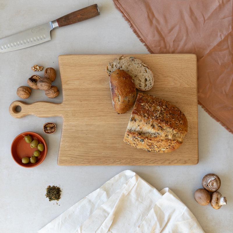 Top view of an oak chopping board against a neutral background. There is a loaf of seeded bread on the board and a selection of foodstuffs around the board including walnuts, mushrooms and olives.