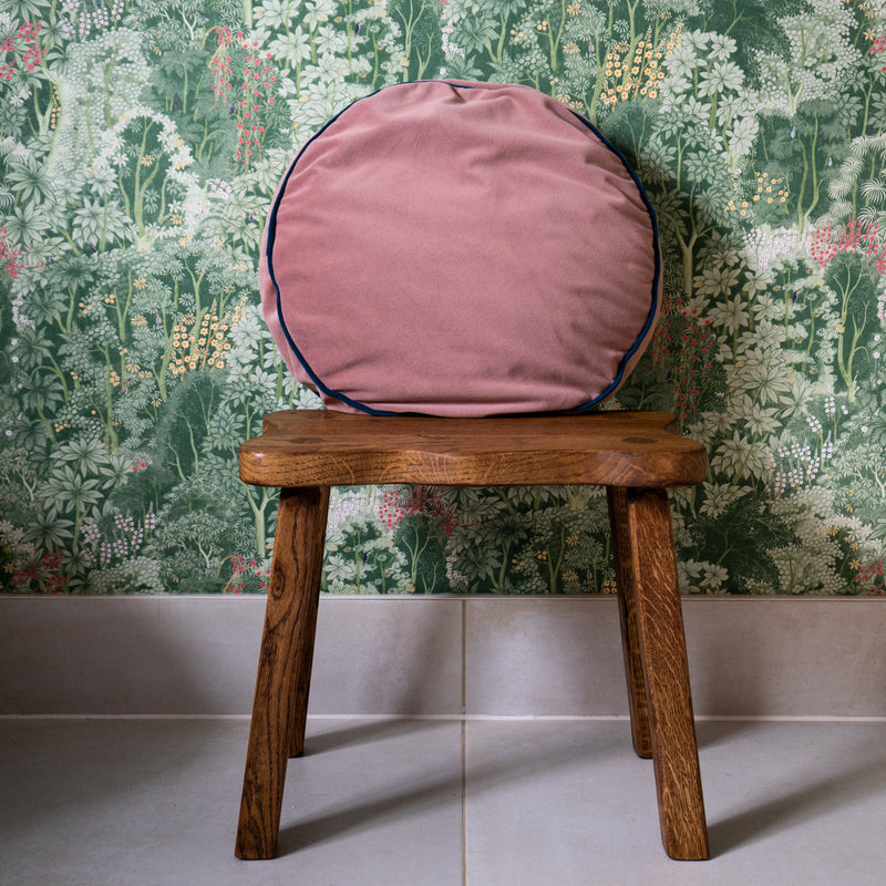 A chunky stool with four legs is placed on a tiled floor against wall with botanical wallpaper. There is a pink, circular cushion placed on top the stool. The top surface of the stool is an irregular wavy shape.