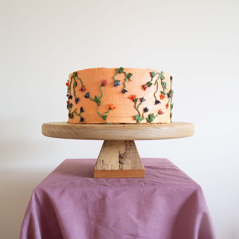 A peach coloured cake with a floral design on top of a wooden cake stand. The base of the cake stand is made from spalted ash and sapele. The stand is on some purple fabric against an off-white wall.