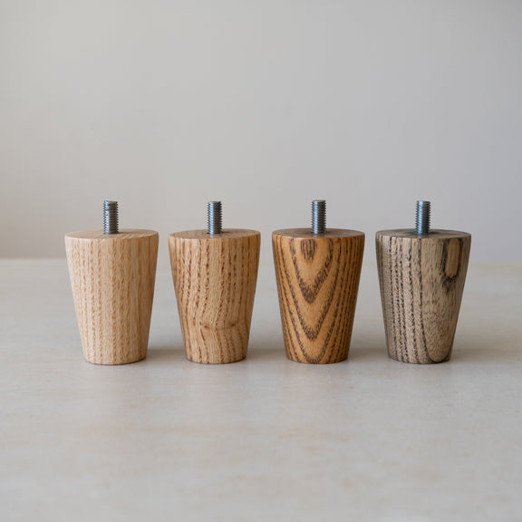 Four tapered ash furniture legs with M8 screw threads against a neutral background. All four legs are in different finishes - Clear, Medium Brown, Warm Dark Brown and Onyx Grey.