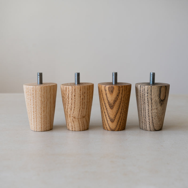 Four tapered ash furniture legs with M8 screw threads against a neutral background. All four legs are in different finishes - Clear, Medium Brown, Warm Dark Brown and Onyx Grey.