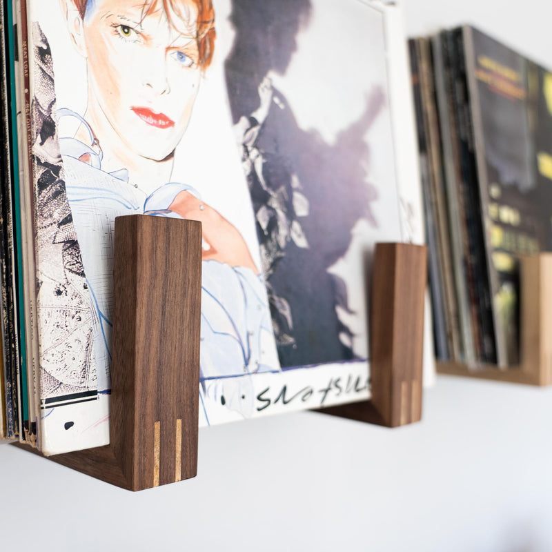 Close up side view of a walnut record rack filled with records. The record at the front is a David Bowie album. An oak rack is blurred out in the background.